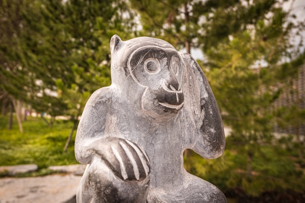 A stone sculpture of a monkey sits amidst lush greenery. The monkey appears to be holding one paw near its ear, as if listening attentively.