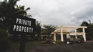 A weathered sign reading 'Private Property' is positioned in the foreground, surrounded by lush tropical plants. In the background, a partially constructed or abandoned white structure is situated under an overcast sky. The ground is scattered with wooden debris and there is a collection of outdoor furniture scattered near the building.