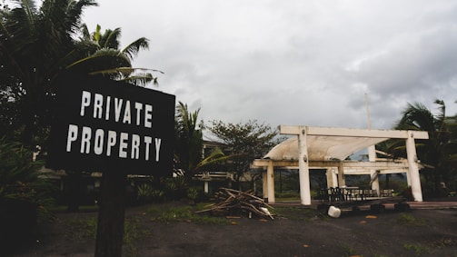 A weathered sign reading 'Private Property' is positioned in the foreground, surrounded by lush tropical plants. In the background, a partially constructed or abandoned white structure is situated under an overcast sky. The ground is scattered with wooden debris and there is a collection of outdoor furniture scattered near the building.