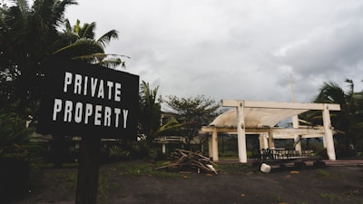 A weathered sign reading 'Private Property' is positioned in the foreground, surrounded by lush tropical plants. In the background, a partially constructed or abandoned white structure is situated under an overcast sky. The ground is scattered with wooden debris and there is a collection of outdoor furniture scattered near the building.