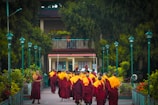 A group of monks wearing orange and burgundy robes walks toward a building surrounded by lush greenery and teal-colored lampposts. The scene is peaceful, situated in a tranquil garden-like setting.