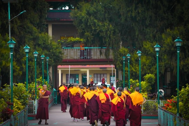 A group of monks wearing orange and burgundy robes walks toward a building surrounded by lush greenery and teal-colored lampposts. The scene is peaceful, situated in a tranquil garden-like setting.