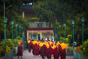 A group of monks wearing orange and burgundy robes walks toward a building surrounded by lush greenery and teal-colored lampposts. The scene is peaceful, situated in a tranquil garden-like setting.