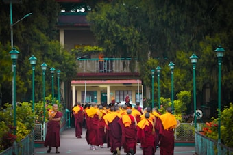 A group of monks wearing orange and burgundy robes walks toward a building surrounded by lush greenery and teal-colored lampposts. The scene is peaceful, situated in a tranquil garden-like setting.