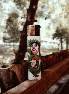 A lighter with artistic illustrations of two people surrounded by leaves is placed on a wooden railing. The background features blurry trees and a diffused light creating a warm ambiance.