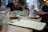 A blurred image of a café setting with a focus on a menu placed on a wooden table. The background features out-of-focus people sitting and conversing, with cups and glasses scattered around.