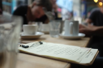 A blurred image of a caf&eacute; setting with a focus on a menu placed on a wooden table. The background features out-of-focus people sitting and conversing, with cups and glasses scattered around.