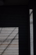 A closed metal roller shutter door with horizontal slats, partially covered in shadow. A small circular sticker can be seen on the right side on a concrete surface next to the door.