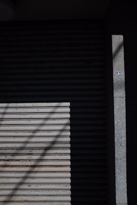 A closed metal roller shutter door with horizontal slats, partially covered in shadow. A small circular sticker can be seen on the right side on a concrete surface next to the door.