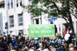 A large group of people gathered outdoors in a public area holding a green protest sign with the phrase 'It's not easy being green' attributed to a character. Bicycles are visible in the background, along with urban buildings and trees.