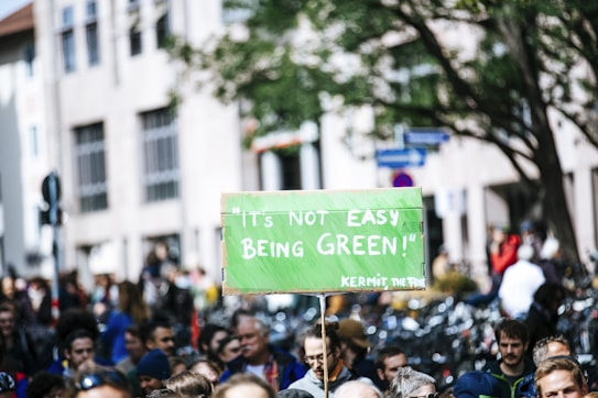 A large group of people gathered outdoors in a public area holding a green protest sign with the phrase 'It's not easy being green' attributed to a character. Bicycles are visible in the background, along with urban buildings and trees.
