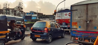 A busy urban street scene features various vehicles including an auto-rickshaw, a taxi with a yellow roof, and a large truck at a traffic intersection. Buildings in the background appear worn down with visible advertising signage on one of them. The sky is cloudy with a hint of sunlight breaking through, casting a warm glow over the scene.