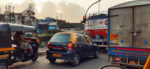 A busy urban street scene features various vehicles including an auto-rickshaw, a taxi with a yellow roof, and a large truck at a traffic intersection. Buildings in the background appear worn down with visible advertising signage on one of them. The sky is cloudy with a hint of sunlight breaking through, casting a warm glow over the scene.
