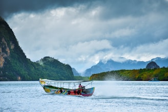 A colorful long-tail boat travels across a calm body of water in a tropical landscape. Lush green hills and mountains surround the water, with mist clinging to their surfaces. Dark clouds loom overhead, adding a dramatic effect to the scene.