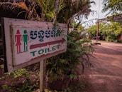 A slightly weathered sign indicating the direction to the toilet, featuring symbols for a male and female. The text is in both English and another language. Surrounding the sign are lush green plants with branches extending around it. The background features an open outdoor setting with a dirt path and more greenery.