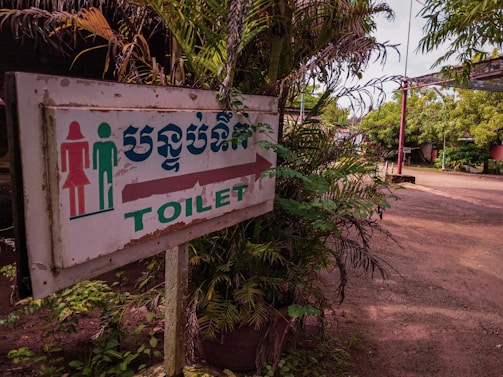 A slightly weathered sign indicating the direction to the toilet, featuring symbols for a male and female. The text is in both English and another language. Surrounding the sign are lush green plants with branches extending around it. The background features an open outdoor setting with a dirt path and more greenery.