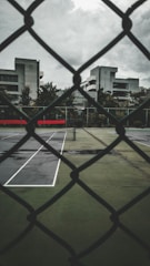 A tennis court is visible through a chain-link fence, with the court's pavement slightly wet from recent rain. The surrounding buildings are modern and utilitarian in design, with overcast skies adding a gloomy atmosphere.