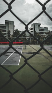 A tennis court is visible through a chain-link fence, with the court's pavement slightly wet from recent rain. The surrounding buildings are modern and utilitarian in design, with overcast skies adding a gloomy atmosphere.