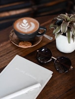 A flatlay composition includes a cup of coffee with latte art on a wooden table, accompanied by a small biscuit. Nearby, a potted plant adds a touch of greenery. Beside the coffee, a pair of round sunglasses rests next to a journal titled 'MY LIFE JOURNAL' and a white pen.