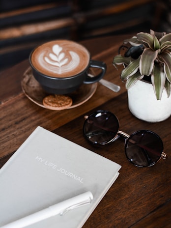 A flatlay composition includes a cup of coffee with latte art on a wooden table, accompanied by a small biscuit. Nearby, a potted plant adds a touch of greenery. Beside the coffee, a pair of round sunglasses rests next to a journal titled 'MY LIFE JOURNAL' and a white pen.