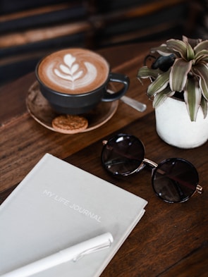 A flatlay composition includes a cup of coffee with latte art on a wooden table, accompanied by a small biscuit. Nearby, a potted plant adds a touch of greenery. Beside the coffee, a pair of round sunglasses rests next to a journal titled 'MY LIFE JOURNAL' and a white pen.