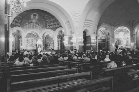 A large congregation of people seated in rows of wooden benches inside a grand church. The interior features arched ceilings and intricate murals depicting religious scenes. Soft lighting creates a serene atmosphere.