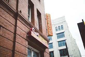 A red-brick building featuring signage for a barbecue restaurant with classic style signage and a neon sign that reads 'BARBECUE' vertically. Adjacent is a white building with multiple windows and an architectural design that contrasts with the rustic look of the brick.