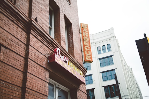 A red-brick building featuring signage for a barbecue restaurant with classic style signage and a neon sign that reads 'BARBECUE' vertically. Adjacent is a white building with multiple windows and an architectural design that contrasts with the rustic look of the brick.