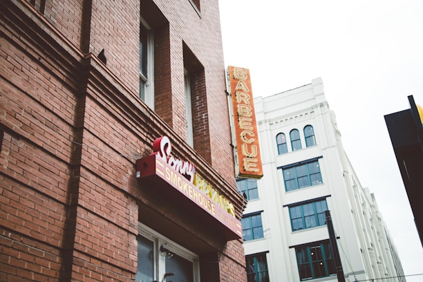 A red-brick building featuring signage for a barbecue restaurant with classic style signage and a neon sign that reads 'BARBECUE' vertically. Adjacent is a white building with multiple windows and an architectural design that contrasts with the rustic look of the brick.