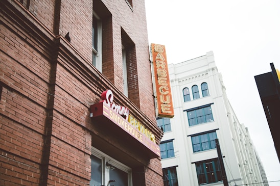A red-brick building featuring signage for a barbecue restaurant with classic style signage and a neon sign that reads 'BARBECUE' vertically. Adjacent is a white building with multiple windows and an architectural design that contrasts with the rustic look of the brick.