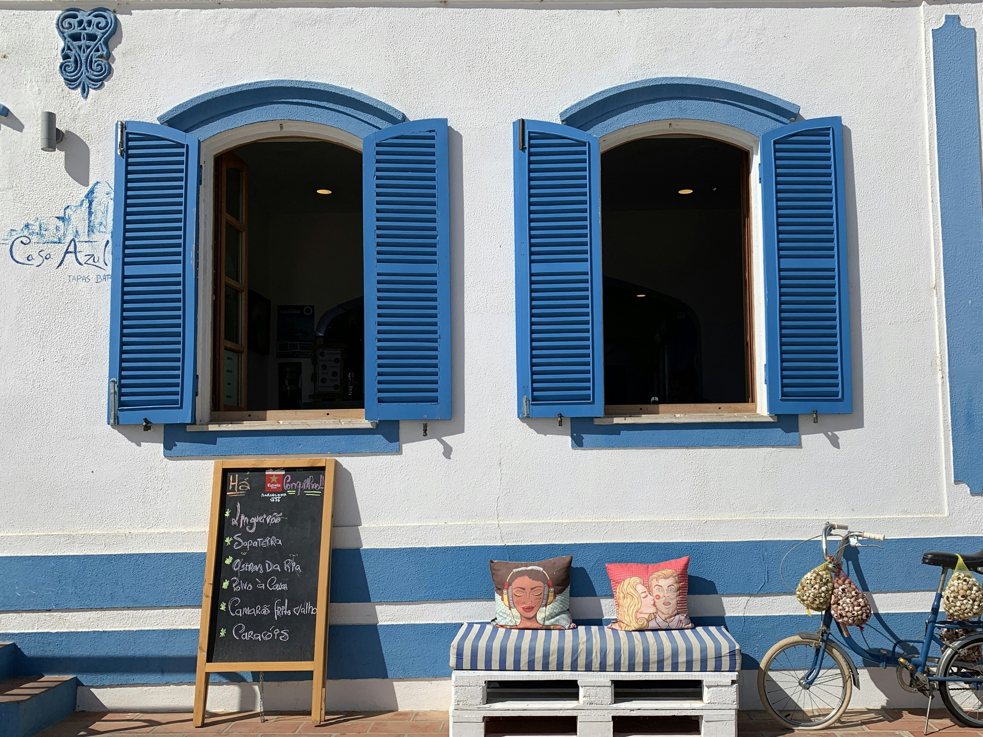 two multicolored throw pillows on bench near bike parking on white and blue wall showing open blue wooden windows, 