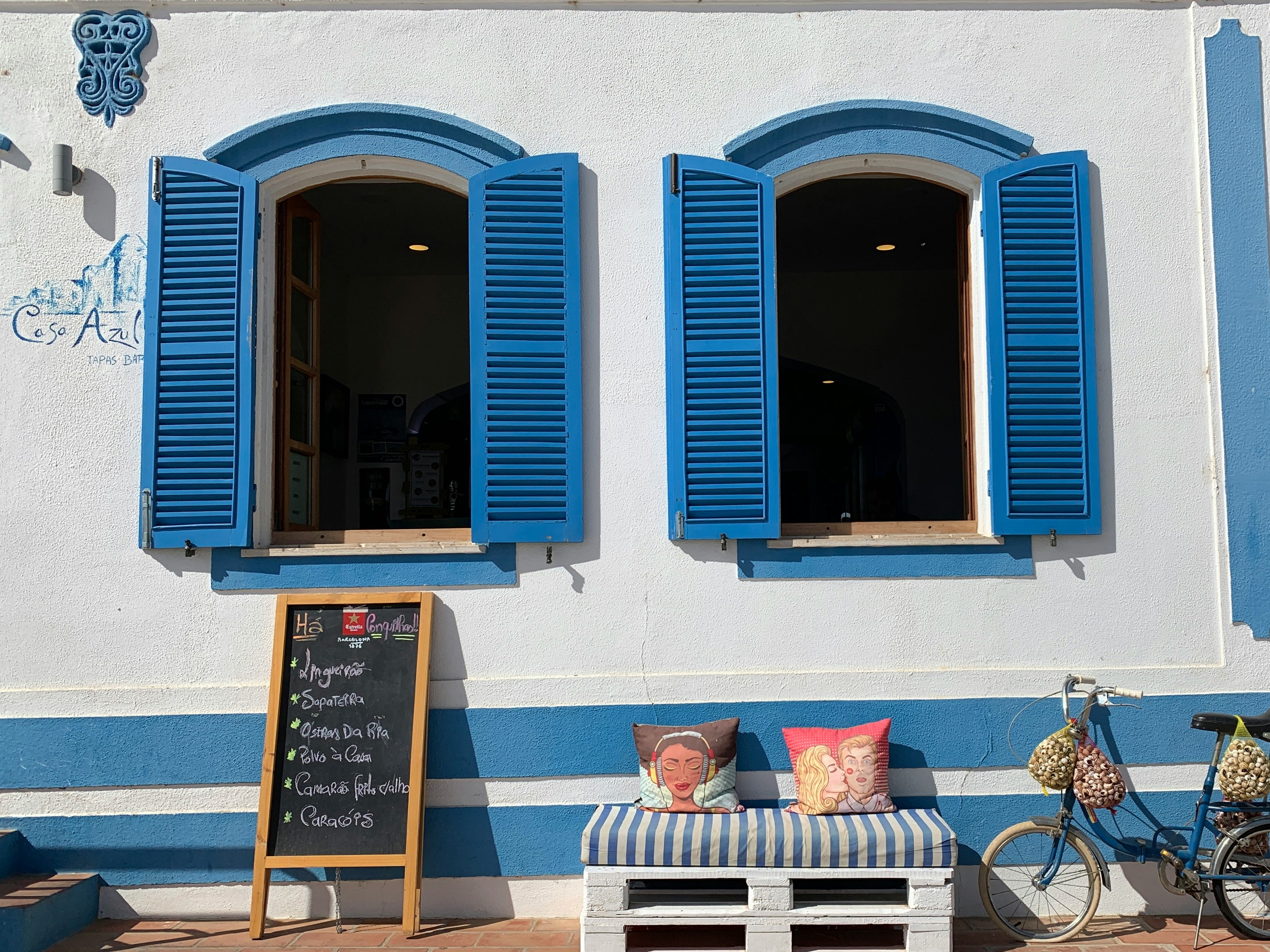 two multicolored throw pillows on bench near bike parking on white and blue wall showing open blue wooden windows