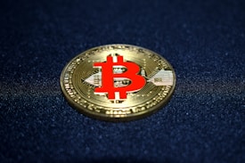 A physical representation of a Bitcoin with a golden color and a prominent red Bitcoin symbol at its center, resting on a dark textured surface.