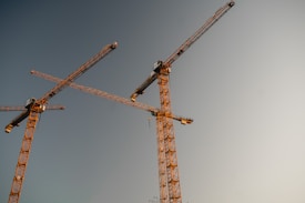 Two large construction cranes are positioned in the foreground against a clear sky during what appears to be sunset or sunrise. The cranes are primarily orange and tower into the sky, indicating construction or development activities. The background is a soft gradient of blues and warm tones.