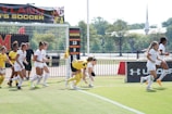 A group of female soccer players from Maryland are running across a soccer field. Some players are in white uniforms with red accents, while a few others wear yellow jerseys. In the background, banners display accolades like NCAA tournaments and achievements. Trees and a distant building are visible beyond the field.