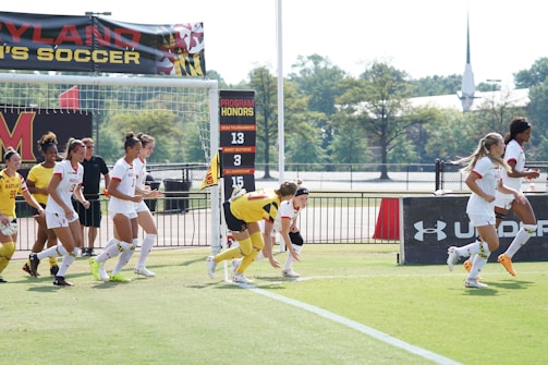 A group of female soccer players from Maryland are running across a soccer field. Some players are in white uniforms with red accents, while a few others wear yellow jerseys. In the background, banners display accolades like NCAA tournaments and achievements. Trees and a distant building are visible beyond the field.