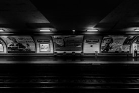 A monochromatic photograph of an underground metro station platform named Vaugirard. Illuminated advertisements cover the walls, and the platform is mostly devoid of people. Empty metal seats are arranged along the platform, and the view includes overhead lighting fixtures.
