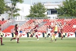 A women's soccer match is underway on a sunny day, with players actively engaged in the game. The players are dressed in maroon and white uniforms, competing on a lush green field surrounded by a stadium with red seats. Spectators can be seen watching the match.