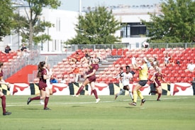 A women's soccer match is underway on a sunny day, with players actively engaged in the game. The players are dressed in maroon and white uniforms, competing on a lush green field surrounded by a stadium with red seats. Spectators can be seen watching the match.