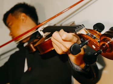 A person is playing a violin, focusing on the hand holding the violin neck and the bow in motion. The violin's strings and tuning pegs are visible in detail. The background is blurred, emphasizing the musician's concentration.