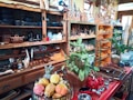 A cozy store filled with handcrafted wooden artifacts, pottery, and decorative figurines. Shelves display an array of clay pots, black kettles, and colorful ceramics along with wooden utensils. Green plants add a touch of nature, and a basket of fresh fruit sits atop a red counter, which also holds an assortment of wooden bowls and red coffee pots.