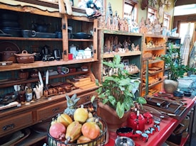 A cozy store filled with handcrafted wooden artifacts, pottery, and decorative figurines. Shelves display an array of clay pots, black kettles, and colorful ceramics along with wooden utensils. Green plants add a touch of nature, and a basket of fresh fruit sits atop a red counter, which also holds an assortment of wooden bowls and red coffee pots.