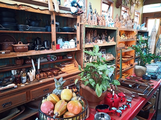 A cozy store filled with handcrafted wooden artifacts, pottery, and decorative figurines. Shelves display an array of clay pots, black kettles, and colorful ceramics along with wooden utensils. Green plants add a touch of nature, and a basket of fresh fruit sits atop a red counter, which also holds an assortment of wooden bowls and red coffee pots.