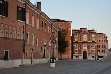 A square in Italy with historic buildings featuring ornate architectural details. The warm-colored facades of the buildings are bathed in the soft light of late afternoon, with a clear sky above. A couple of lampposts and some greenery add charm to the scene.