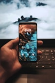 A hand holding a smartphone displaying a night-themed wallpaper with app icons. In the background, there is a laptop with a cloudy sky on the screen.