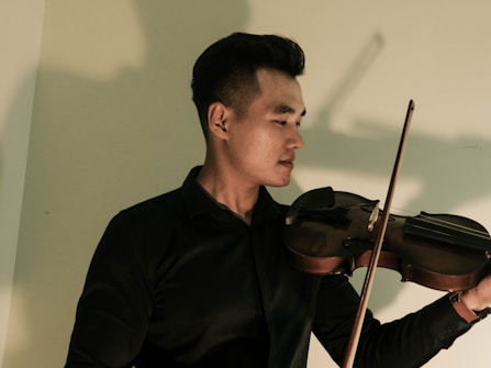 A person in a dark shirt is playing a violin, standing in front of a light-colored wall. The lighting casts a shadow on the wall behind the person, adding depth to the image. The violin is being held gracefully, and the person's expression appears focused and serene.