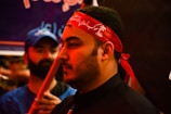 The image shows a close-up of a man wearing a red headband with white script. His expression is calm and focused. In the background, another person in a blue shirt and cap is visible, slightly out of focus.