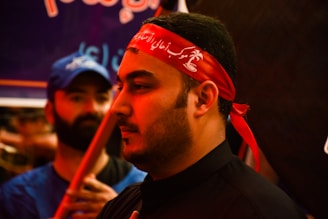 The image shows a close-up of a man wearing a red headband with white script. His expression is calm and focused. In the background, another person in a blue shirt and cap is visible, slightly out of focus.