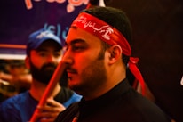 The image shows a close-up of a man wearing a red headband with white script. His expression is calm and focused. In the background, another person in a blue shirt and cap is visible, slightly out of focus.