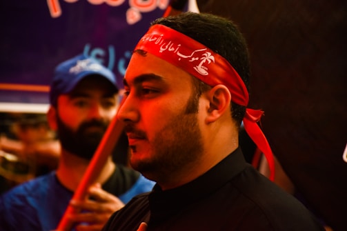The image shows a close-up of a man wearing a red headband with white script. His expression is calm and focused. In the background, another person in a blue shirt and cap is visible, slightly out of focus.
